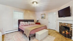 A bedroom with a bed, a bench, a fireplace, a television, and a door. - Lands End House at Cape Porpoise with Hot Tub (Kennebunkport)