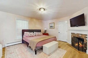 A bedroom with a bed, a bench, a fireplace, a television, and a door. - Lands End House at Cape Porpoise with Hot Tub (Kennebunkport)