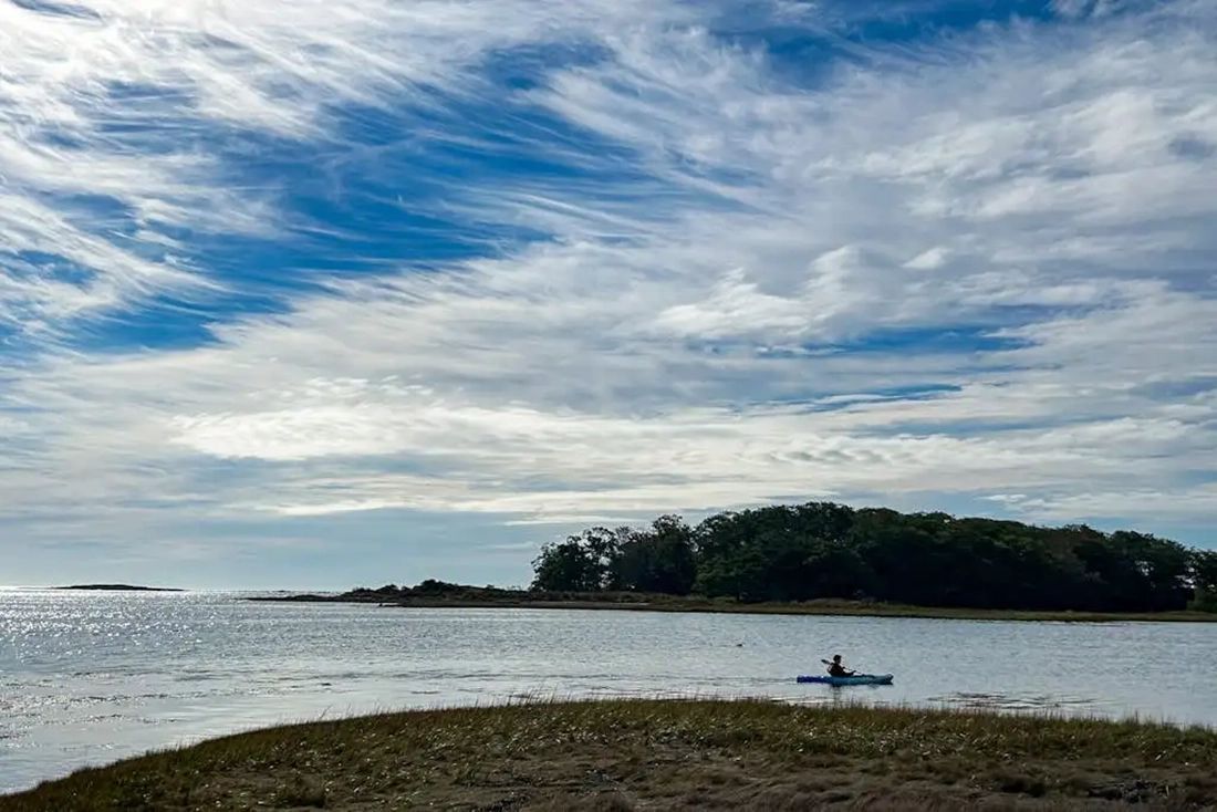 A calm body of water with a single person kayaking, surrounded by a natural landscape and a clear sky.
