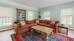 A living room with a sectional sofa, a coffee table, a bookshelf, and a guitar. - Lands End House at Cape Porpoise with Hot Tub (Kennebunkport)