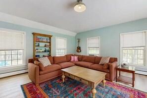 A living room with a sectional sofa, a coffee table, a bookshelf, and a guitar. - Lands End House at Cape Porpoise with Hot Tub (Kennebunkport)