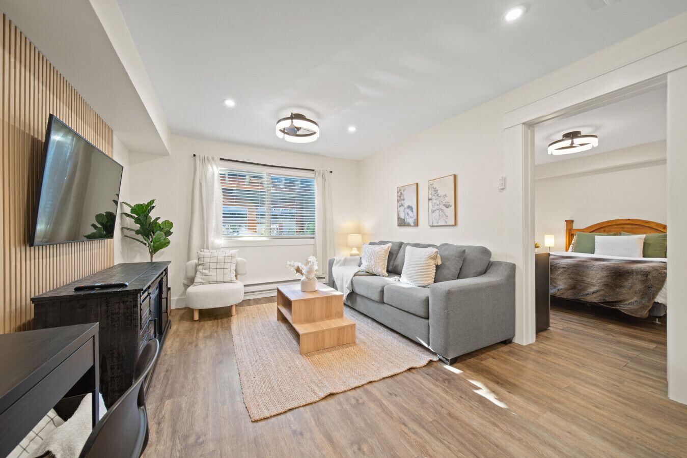 Un salon moderne avec un canapé gris, une table basse en bois, un téléviseur et une fenêtre avec des stores.