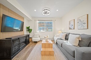 A modern living room with a flat-screen TV mounted on a wooden panel wall, a grey sofa, a wooden coffee table, and a white armchair. - The Lakeside Hideaway (Whistler)