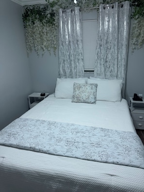 A bedroom with a bed, a nightstand, a bedside table, and a window with curtains. - The Enchanted Garden Suite (Pinellas Park)