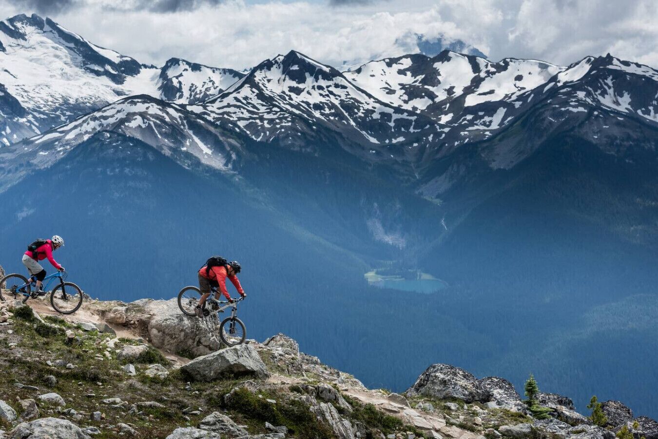 Two mountain bikers on a rocky trail with snow-capped mountains in the background.