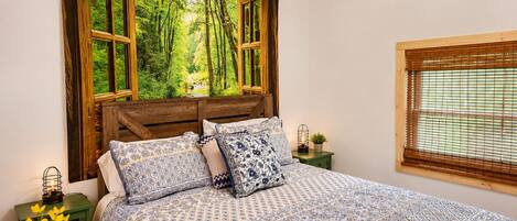 A bedroom with a large bed, a window with a view of a forest, a green nightstand, and a lamp.