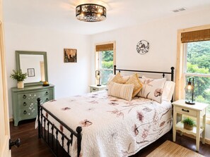 A bedroom with a bed, a dresser, a nightstand, a lamp, a window with a view, and a wall decoration.
