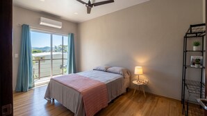 Un dormitorio con cama, lámpara de pie, estante con libros y plantas, y vista al exterior por una ventana.