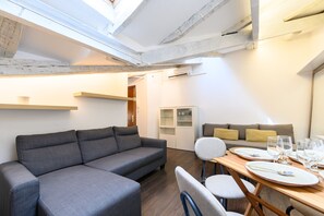 Ein zeitgemäßer Wohnraum mit grauem Sofa, einem Esstisch für vier Personen und einer kleinen Sitzecke mit Kissen.