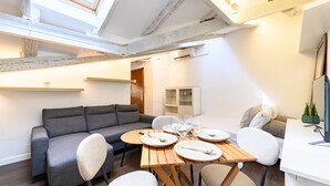 A modern living room with a sofa, dining table, and chairs. There is a window with a view of the sky. - (FER3452) 1 bedroom penthouse in Moncloa (Madrid)
