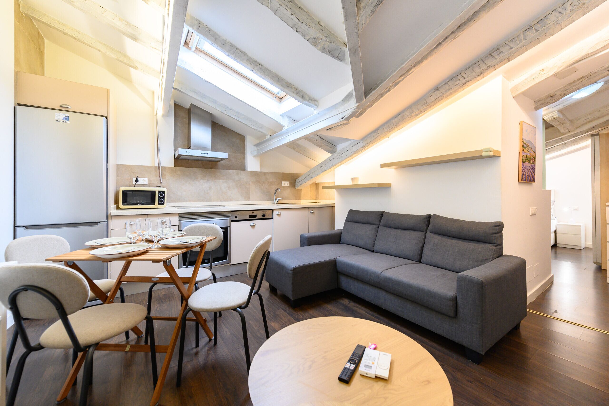 A modern kitchen with a microwave, stove, and sink. There is a dining area with a table and chairs. A gray sofa is in the living area.