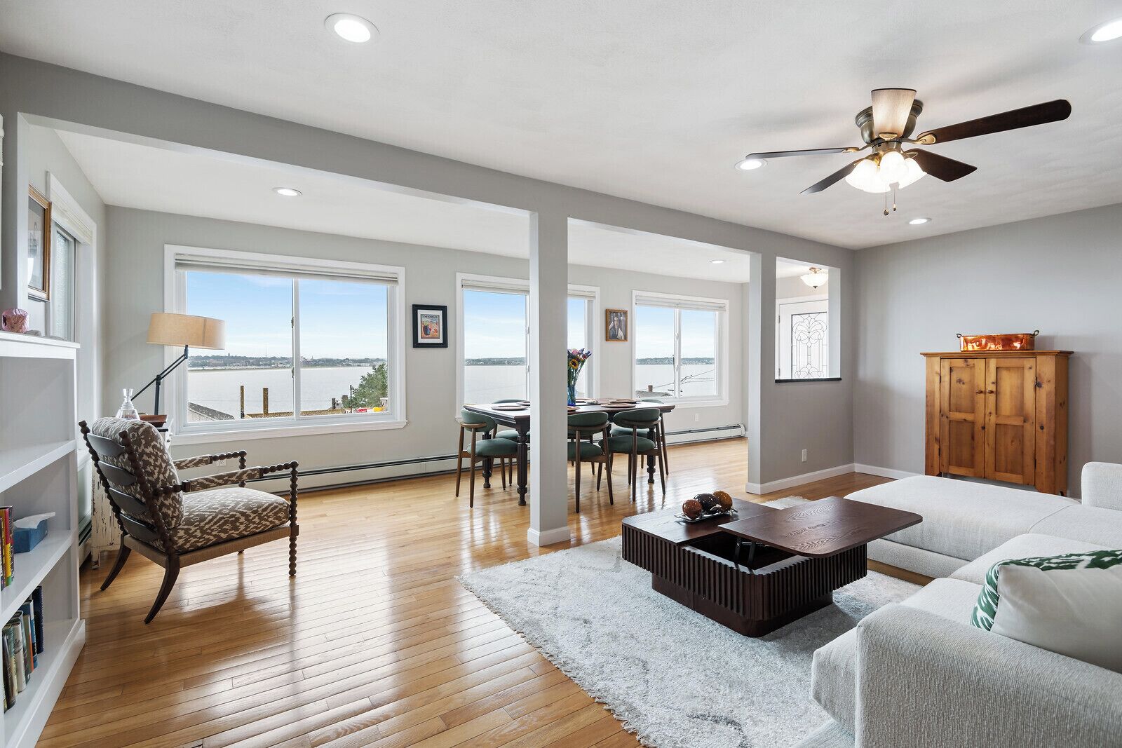 Un vaste salon avec vue sur l’eau, doté d’un ventilateur de plafond et comprenant un coin repas avec une table et des chaises.