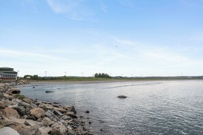 A coastal scene with a building on the left, a rocky shoreline, and a clear sky. - The Nahant Perch: ocean view, walk to beach (Nahant)