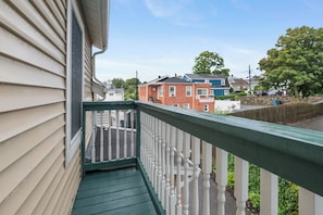 A residential balcony with a green railing, overlooking a street with colorful houses and a tree. - The Nahant Perch: ocean view, walk to beach (Nahant)