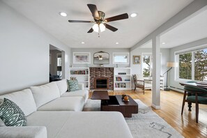 A modern living room with a fireplace, a ceiling fan, and a large window. - The Nahant Perch: ocean view, walk to beach (Nahant)