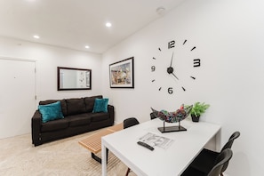 A modern living room with a white wall, a large clock, a sofa with teal cushions, a coffee table, and a potted plant. - SoFi Ocean Drive Villa | Direct Beach Access (Miami)