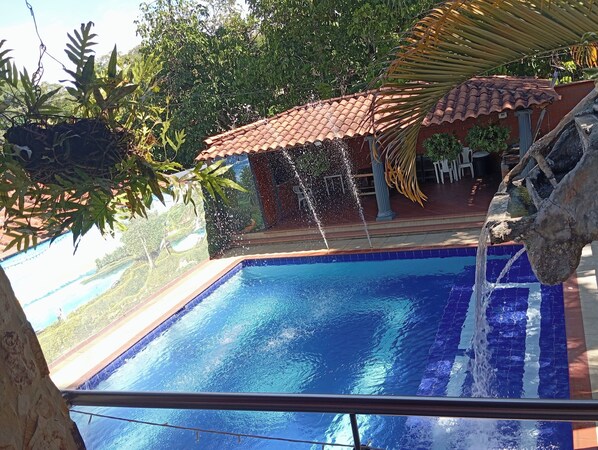 A swimming pool with a water feature, surrounded by greenery and a tiled roof structure.