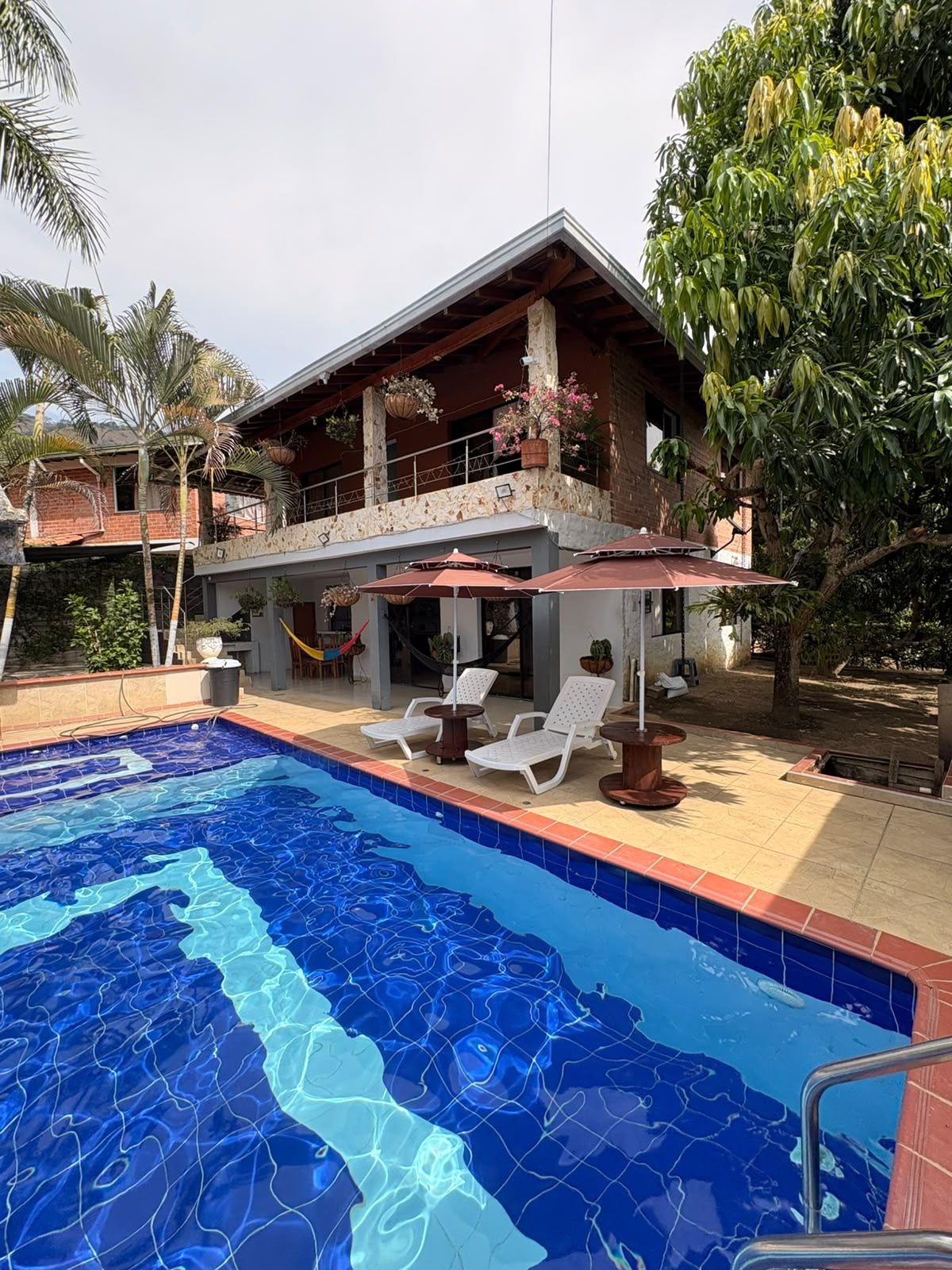 Una casa de dos pisos con piscina, mobiliario exterior y balcón con plantas colgantes.