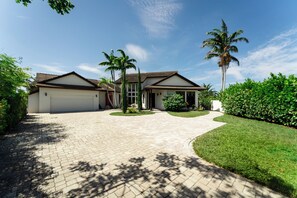 A two-story house with a driveway leading to a garage, surrounded by palm trees and a well-maintained lawn. - Waterfront Luxe Smart Home | Pool | Gym | Spa (Lantana)