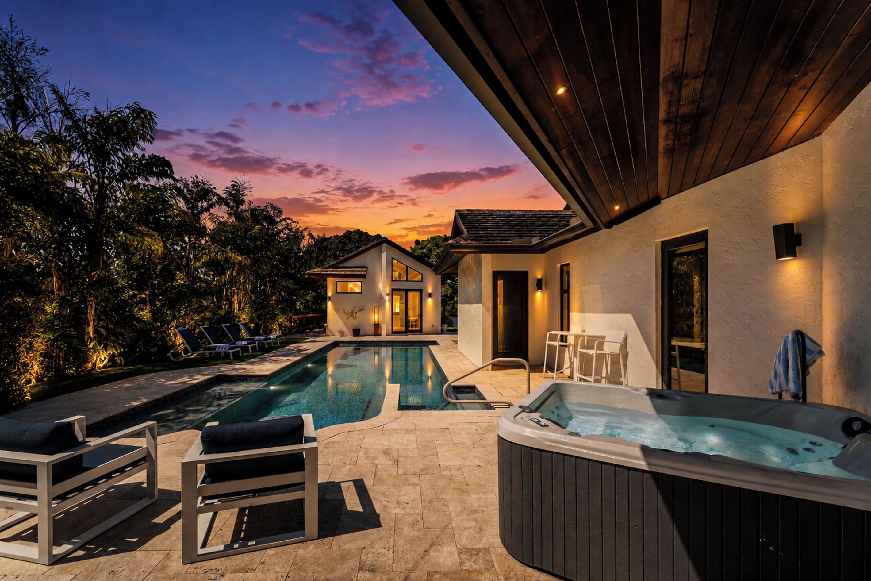 A modern outdoor pool area with lounge chairs, a hot tub, and a house with a patio.