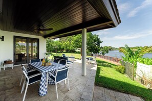 An outdoor patio with a covered seating area, a table with a checkered tablecloth, and a view of a lake. - Waterfront Luxe Smart Home | Pool | Gym | Spa (Lantana)