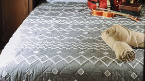 Ein Bett mit einem gemusterten Überwurf, ein Kopfkissen und zusammengerollte Handtücher. Darüber befinden sich Holzschränke und auf dem Boden steht eine Gitarre.