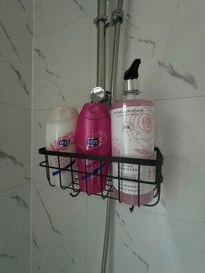 A bathroom shelf with toiletries including a pink bottle of shampoo, a white bottle of conditioner, and a large bottle of perfume.