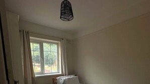 Una habitación pequeña con cama individual, cabecera de madera, ventana con cortinas y una lámpara colgante.