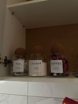 Tres pots de vidre amb etiquetes a una estant, que contenen te, sucre i cafè.