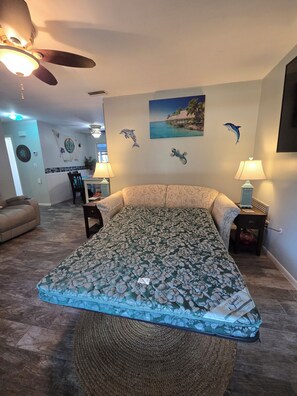 Ein Schlafzimmer mit blumigem Bettüberwurf, Deckenventilator, Wandfernseher und Blick auf das Wohnzimmer.