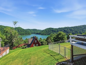 Una casa con techo rojo, patio cercado y vista a un lago y colinas boscosas.