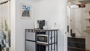 Un área de cocina con cafetera, microondas y refrigerador. Hay un cuadro en la pared y una escalera que sube a un nivel superior.