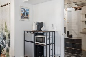 Eine Küchenzeile mit Kaffeemaschine, Mikrowelle und Kühlschrank. An der Wand hängt ein gerahmtes Bild und eine Treppe führt zu einer oberen Ebene.