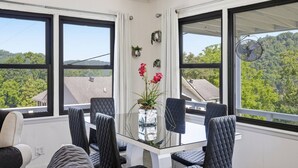 Una habitación con una mesa de vidrio rodeada de sillas, amplias ventanas que dan a vistas de árboles y edificios, y un ventilador de techo.