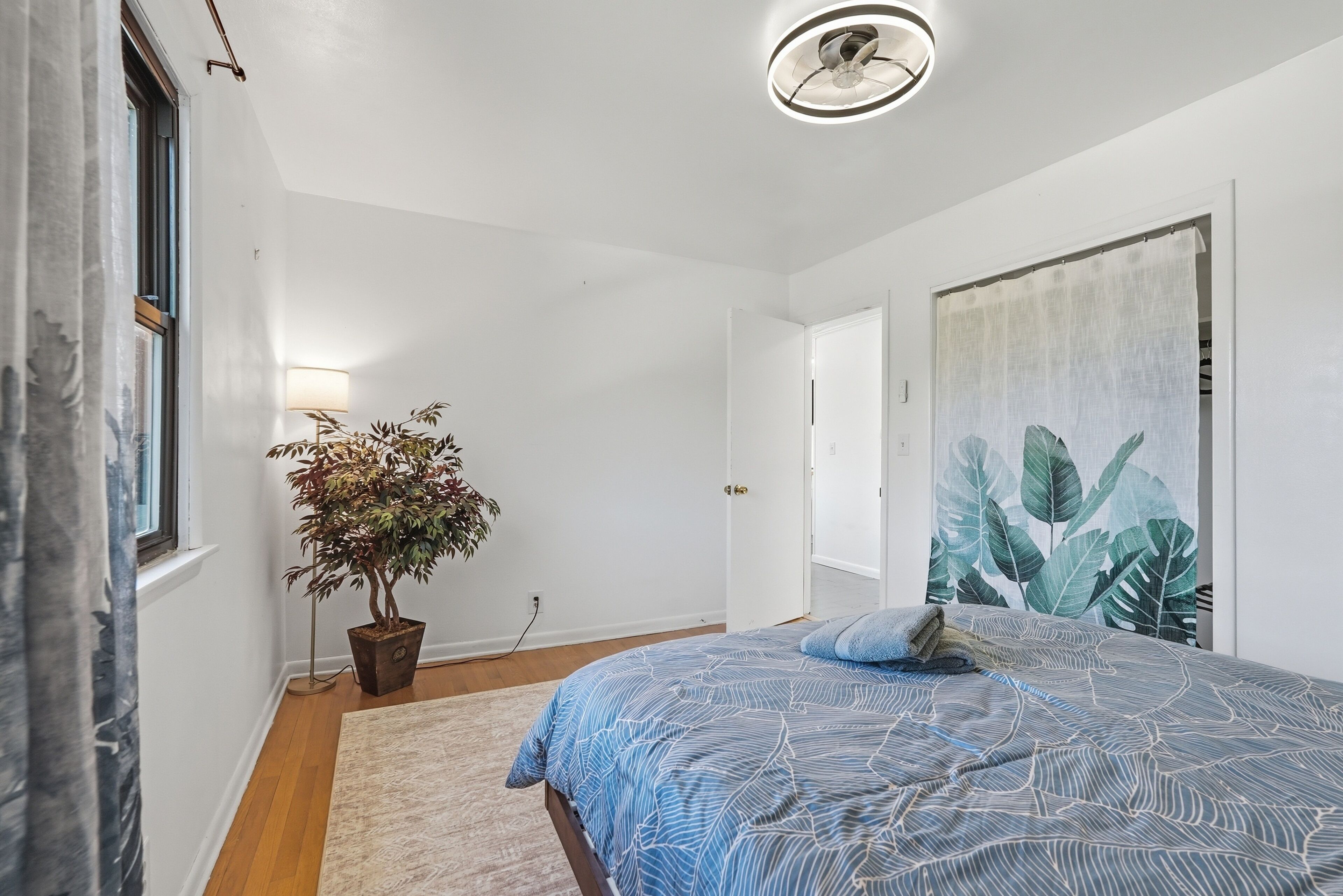 A bedroom with a bed, a potted plant, a wall-mounted lamp, and a large leaf-patterned painting.