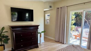 A living room with a dark wooden dresser, a flat-screen TV, a beige wall, and a view of an outdoor patio. - Private retreat w hot tub, sauna & cold plunge steps from La Jolla Shores Beach (San Diego)