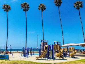 Une aire de jeux de plage avec des toboggans et des structures d’escalade, des palmiers et un ciel bleu azur.