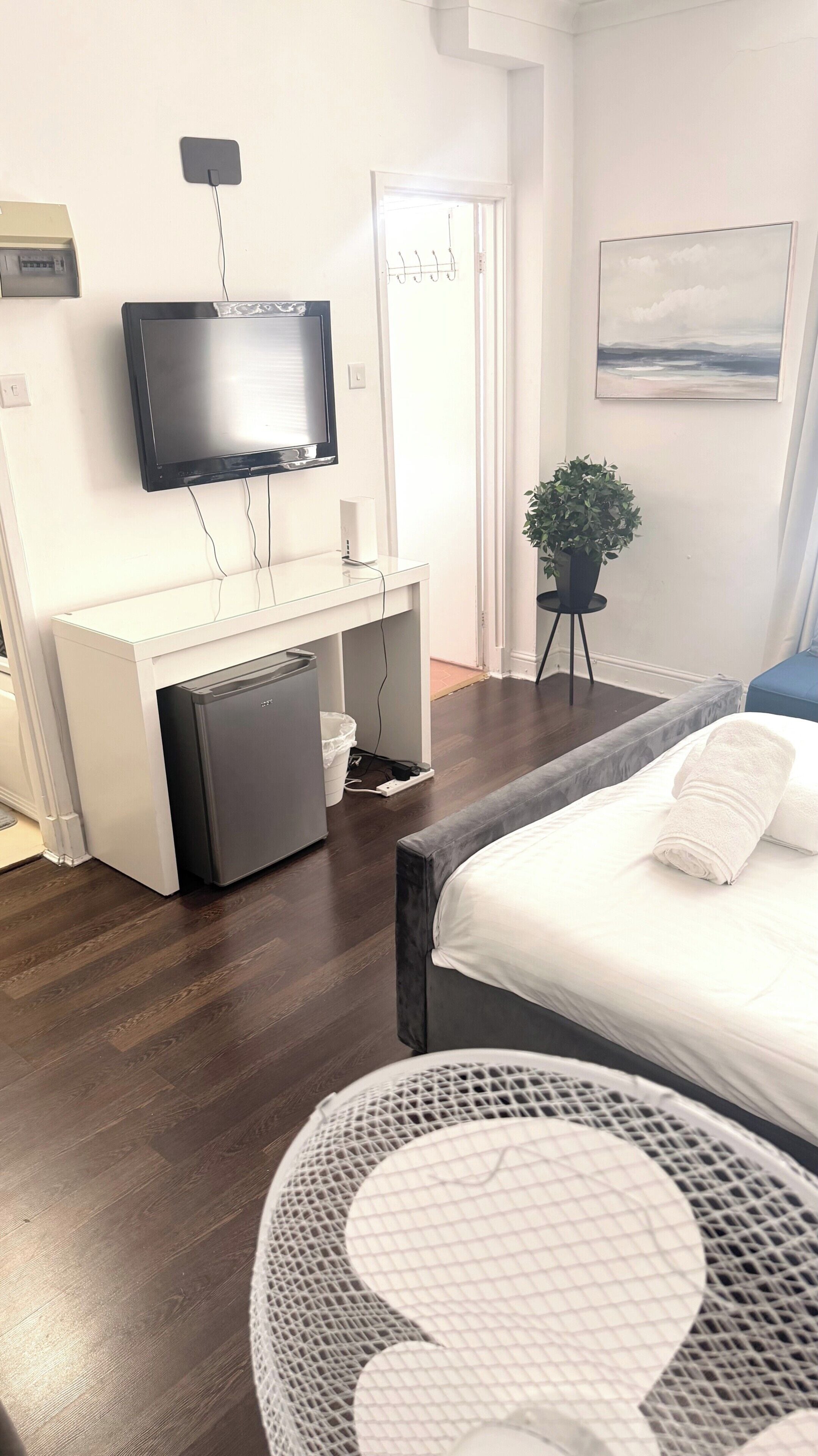 A hotel room with a bed, a desk with a computer, a television, a small refrigerator, a plant, and a painting on the wall.