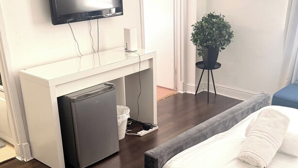 A hotel room with a bed, a desk with a computer, a television, a small refrigerator, a plant, and a painting on the wall. - Cozy London Apartment With City Views (London)