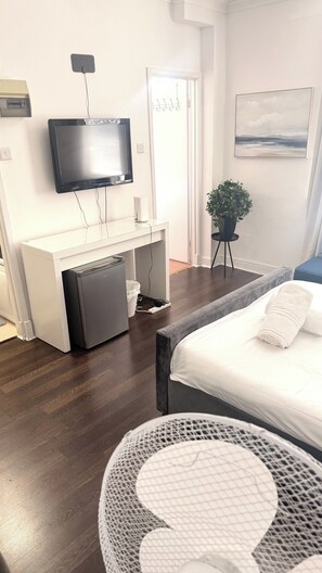 A hotel room with a bed, a desk with a computer, a television, a small refrigerator, a plant, and a painting on the wall. - Cozy London Apartment With City Views (London)