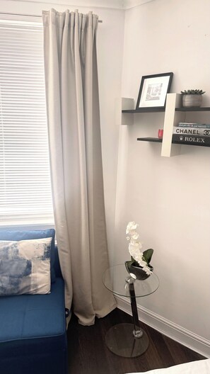 A corner room with a blue sofa, a glass table with an orchid, a white curtain, and a shelf with books and a plant. - Cozy London Apartment With City Views (London)