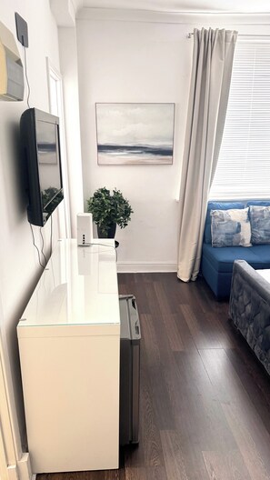 A modern living room with a white desk, a mounted flat-screen TV, a potted plant, a blue sofa with cushions, and a large window with curtains. - Cozy London Apartment With City Views (London)