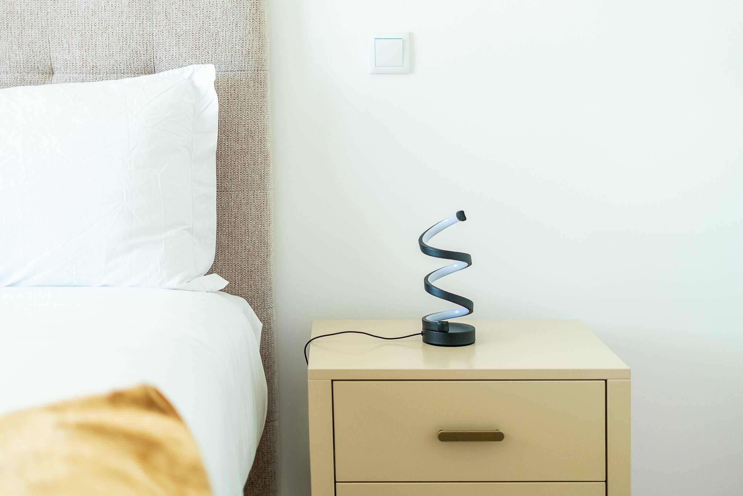 Un letto con biancheria bianca, una testata beige e un comodino con una lampada.