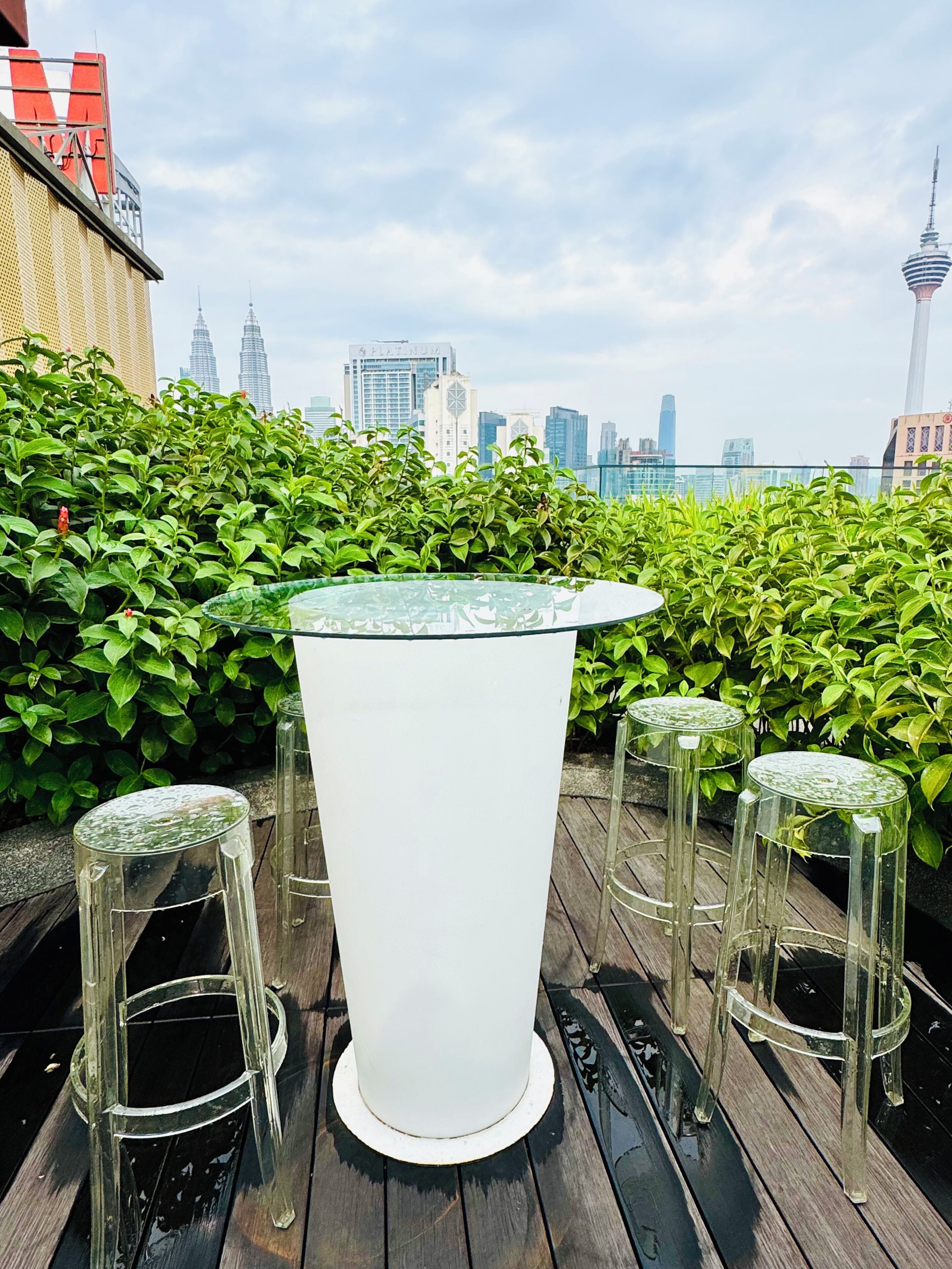 A round table with stools on a wooden deck, surrounded by green foliage, with a cityscape in the background.