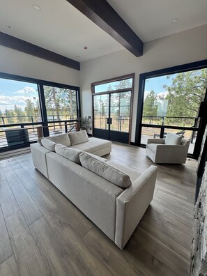 Uma sala de estar espaçosa com um sofá seccional grande, um sofá menor e uma mesa de centro. O espaço possui janelas amplas com vista para o exterior.