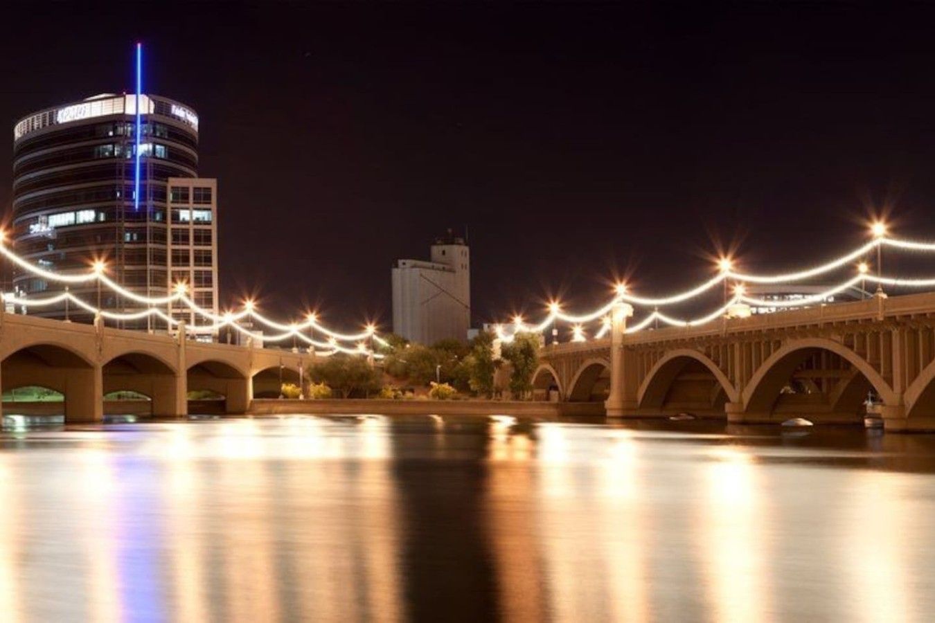 A bridge with arches and string lights, a body of water in the foreground, and a building with a blue-lit top in the background.