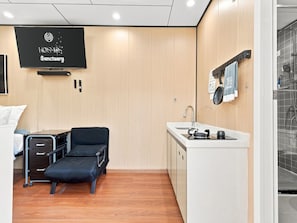 Een compacte hotelkamer met een bed, een bureau, een zwarte stoel, een kleine kitchenette en een televisie waarop 'KOSMOS Sanctuary' staat.