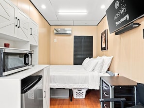 A compact room with a bed, a microwave, a small table, and a TV mounted on the wall. - The Sun Residence #3 at Kosmos Sanctuary (Phoenix)