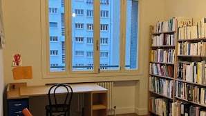 Ein Zimmer mit Schreibtisch, Stuhl, Bücherregal und einem Fenster mit Blick auf Gebäude.