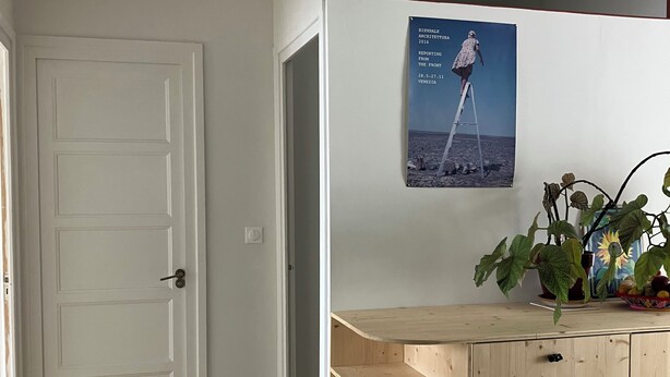 Ein Zimmer mit weißer Tür, einem Holztisch, einem Poster an der Wand und einer Pflanze.